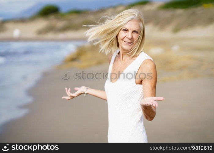 Smiling mature female opening her arms on a tropical beach. Elderly woman enjoying her retirement walking on the beach.. Happy mature woman walking on the beach, spending her leisure time, enjoying her free time