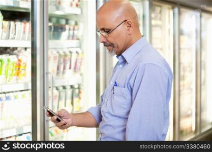 Smiling mature man looking at mobile phone while standing in front of refrigerator in supermarket