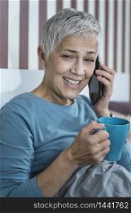 Smiling mature woman sitting in bedroom, drinking tea and talking over the phone before sleep.. Smiling Mature Woman Drinking Tea and Talking Over the Phone Before Sleep