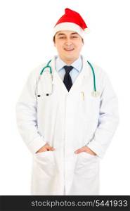 Smiling medical doctor in Santa hat keeping his hands in pockets isolated on white&#xA;