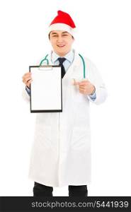 Smiling medical doctor in Santa hat pointing finger at blank clipboard isolated on white&#xA;