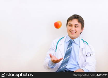 Smiling medical doctor sitting on floor and throwing up apple