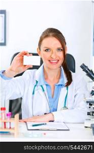 Smiling medical doctor woman sitting at office table and holding blank business card&#xA;