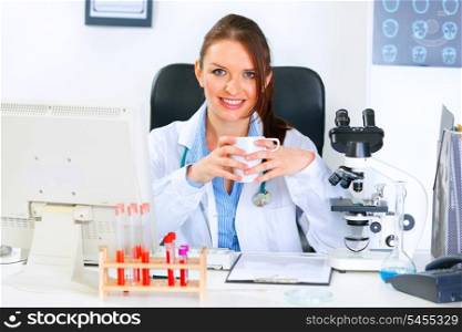 Smiling medical doctor woman sitting at office table and holding cup in hands&#xA;