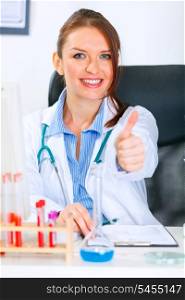 Smiling medical doctor woman sitting at office table and showing thumbs up gesture