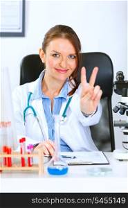 Smiling medical doctor woman sitting at office table and showing victory gesture&#xA;
