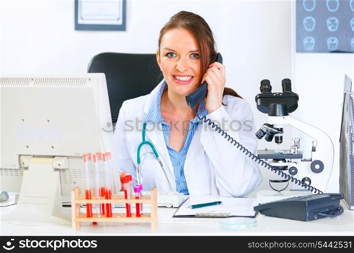 Smiling medical doctor woman sitting at office table and talking on phone&#xA;