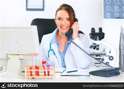 Smiling medical doctor woman sitting at office table and talking on phone&#xA;