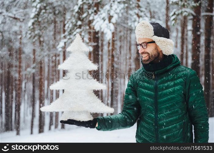 Smiling middle aged male looks with happy expression at artificial white small fir tree, going to celebrate New Year`s holidays, spends morning on fresh air in forest during snowy weather