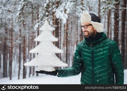 Smiling middle aged male looks with happy expression at artificial white small fir tree, going to celebrate New Year`s holidays, spends morning on fresh air in forest during snowy weather
