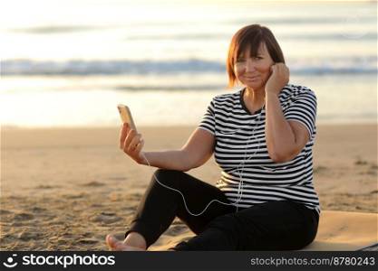 smiling middle ages woman in sportswear uses a smartphone and listens to music with headphones while sitting on a mat on the beach at sunrise. active fitness lifestyle, outdoor workout. smiling middle ages woman in sportswear uses a smartphone and listens to music with headphones while sitting on a mat on the beach at sunrise. active fitness lifestyle, outdoor workout.
