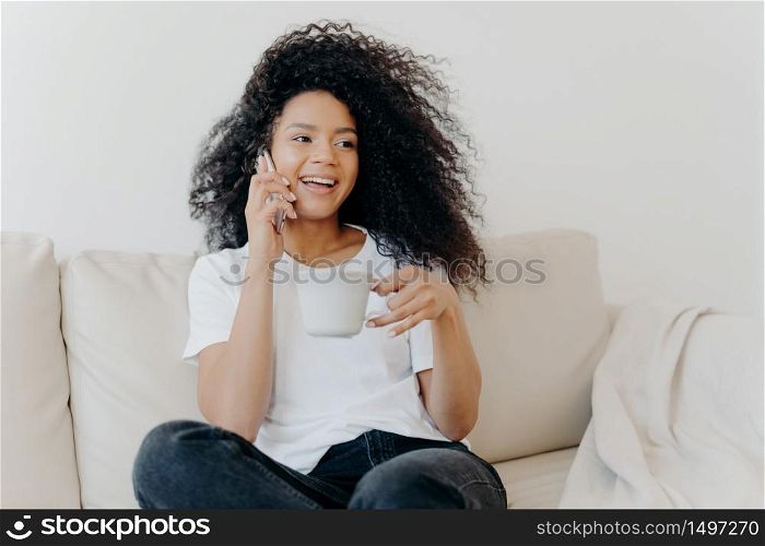 Smiling millennial girl with Afro hair, enjoys cellphone conversation with hot drink in cup, has healthy skin, dressed casually, sits on white sofa at home in modern apartment, orders delivery