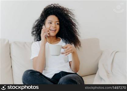 Smiling millennial girl with Afro hair, enjoys cellphone conversation with hot drink in cup, has healthy skin, dressed casually, sits on white sofa at home in modern apartment, orders delivery