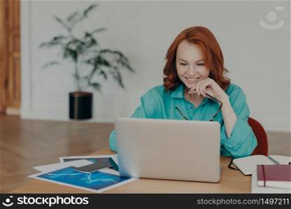 Smiling millennial woman with red hair works or studies on modern laptop computer, browses internet, has video conference, gives online consultation to clients, gets job promotion or nice offer