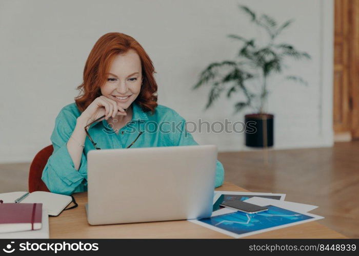 Smiling millennial woman with red hair works or studies on modern laptop computer, browses internet, has video conference, gives online consultation to clients, gets job promotion or nice offer