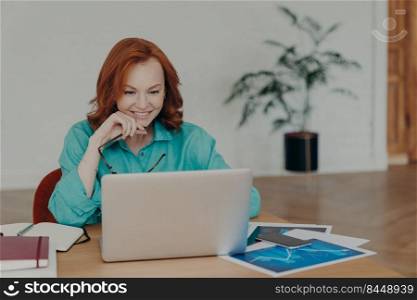Smiling millennial woman with red hair works or studies on modern laptop computer, browses internet, has video conference, gives online consultation to clients, gets job promotion or nice offer