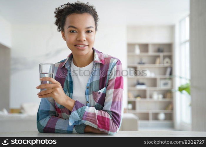 Smiling mixed race girl holding glass of pure water, sitting at home. Happy pretty biracial young woman holds fresh mineral water, looking at camera. Healthy lifestyle, daily routine concept.. Young mixed race girl holding glass of pure water, sitting at home. Healthy lifestyle, self care