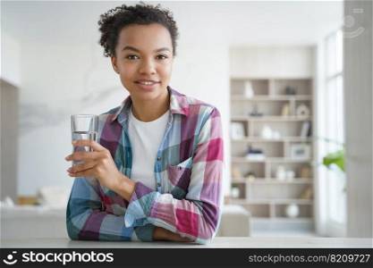 Smiling mixed race girl holding glass of pure water, sitting at home. Happy pretty biracial young woman holds fresh mineral water, looking at camera. Healthy lifestyle, daily routine concept.. Young mixed race girl holding glass of pure water, sitting at home. Healthy lifestyle, self care