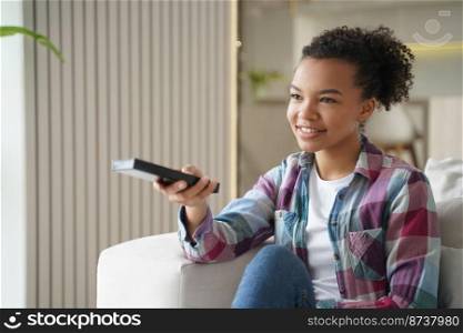 Smiling mixed race girl holding tv remote control, changing channels, resting on couch at home, happy young woman watching television, movie or modern series, spending leisure time on lazy weekend.. Smiling mixed race girl changing TV channels, watching television series, resting on couch at home