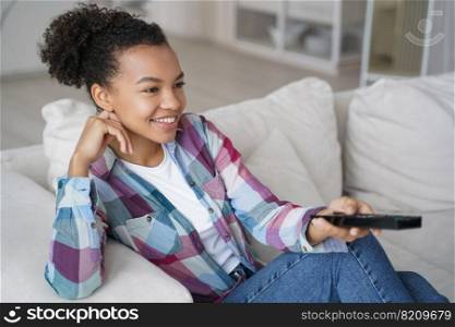 Smiling mixed race teen girl watching television series, sitting on sofa at home. Happy biracial young woman changes TV channel or sound with remote control, relaxing on cozy sofa on weekend.. Smiling mixed race teen girl watching television series, changes TV channel, sitting on sofa at home