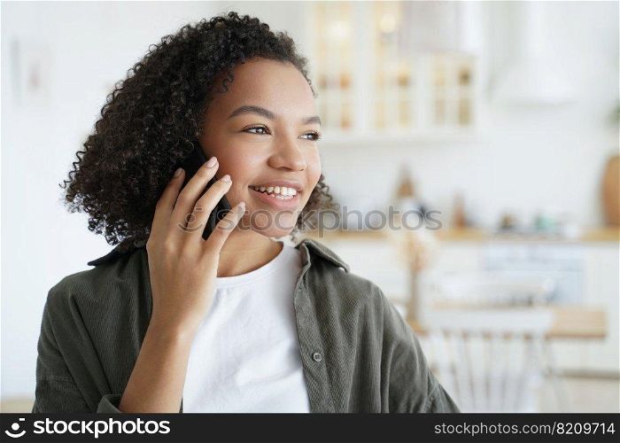 Smiling mixed race young girl answers call, talking on phone, enjoying pleasant mobile conversation at home. Friendly biracial teen lady got good news speaking, chatting by smartphone.. Happy mixed race girl answers call, talking on phone, enjoy pleasant mobile conversation at home