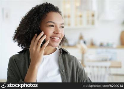 Smiling mixed race young girl answers call, talking on phone, enjoying pleasant mobile conversation at home. Friendly biracial teen lady got good news speaking, chatting by smartphone.. Happy mixed race girl answers call, talking on phone, enjoy pleasant mobile conversation at home