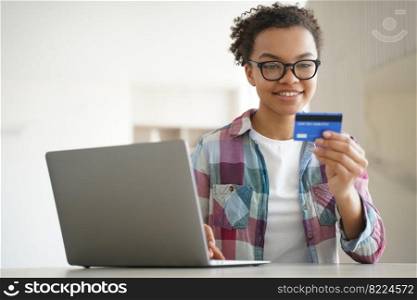 Smiling mixed race young girl holding credit card, using online banking services on laptop indoors. Happy teen lady in glasses enters bank account details on computer at home. E-banking, e-commerce.. Smiling mixed race young girl holding credit card uses online banking services on laptop. E-banking