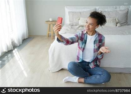 Smiling mixed race young girl talking online by video call, sitting on the floor in bedroom. Happy teenager blogger holding smartphone, chatting with followers in live air in social network at home.. Smiling mixed race young girl blogger talking online by video call, sitting on the floor in bedroom