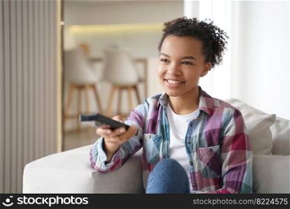 Smiling mixed race young girl watching television series changes TV channels with remote control, sitting on sofa at home. Happy teen lady watches movie, rests on couch, enjoying leisure time.. Smiling mixed race teen girl watching television series changes TV channels sitting on couch at home