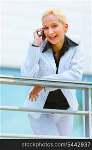 Smiling modern business woman leaning on railing at office building and talking on mobile&#xA;