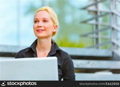 Smiling modern business woman sitting on stairs at office building and using laptop&#xA;