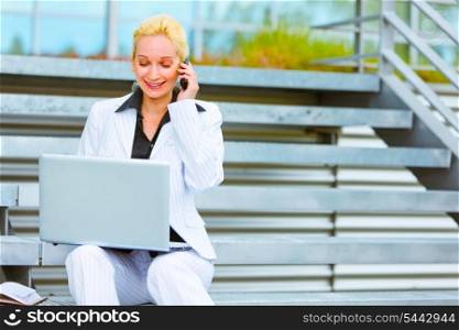 Smiling modern business woman sitting on stairs talking on mobile and looking in laptop &#xA;
