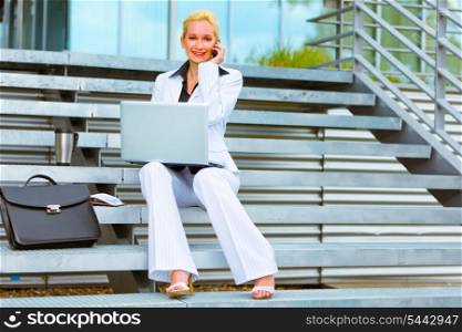 Smiling modern business woman sitting on stairs with laptop and talking on cell phone&#xA;