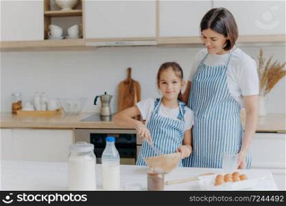 Smiling mother and daughter prepare tasty cookies, girl whisks ingredients in bowl with beater, helps mum at kitchen, prepare festive dinner, modern home interior. Family, cooking, lifestyle concept
