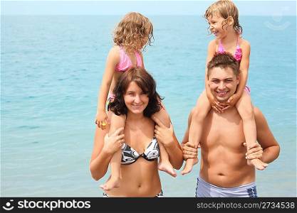 smiling mother and father near water with two little girls sitting on their necks.