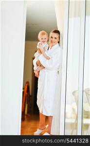 Smiling mother in bathrobe with baby looking out from window