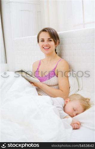 Smiling mother in bed reading book while baby sleeping