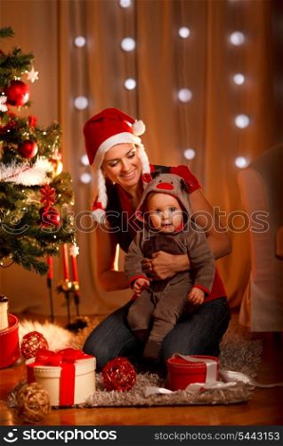 Smiling mother with lovely baby sitting near Christmas tree&#xA;