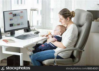 Smiling mother working at home office and feeding her baby from bottle