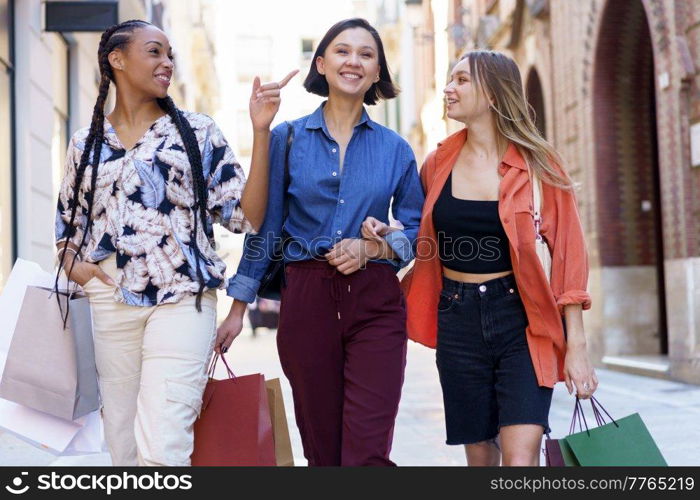 Smiling multiracial female friends in stylish wear with colorful shopping bags walking on sidewalk near buildings after shopping on street. Cheerful diverse women with shopping bags