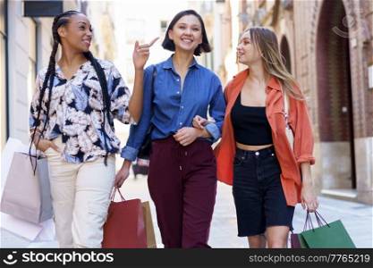 Smiling multiracial female friends in stylish wear with colorful shopping bags walking on sidewalk near buildings after shopping on street. Cheerful diverse women with shopping bags