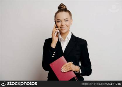 Smiling office worker in black suit, having phone call, holding red notebook, hears good news, isolated on grey background.
