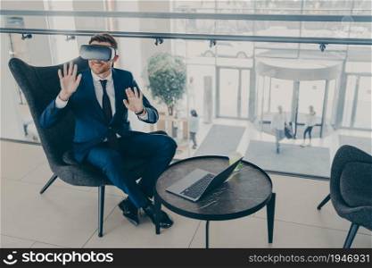 Smiling office worker in formal suit sitting in company center lobby with laptop and VR headset goggles, testing new virtual reality business digital project or app. Innovative technology at work. Smiling office worker sitting in company center lobby with laptop and VR headset goggles