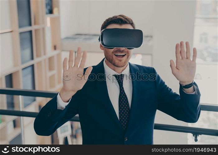 Smiling office worker in suit in 3d goggles enjoying cyberpace experience with raised arms, surprised excited businessman gesturing and touching object in virtual reality, using VR headset. Smiling office worker in suit in 3d goggles enjoying cyberpace experience with raised arms