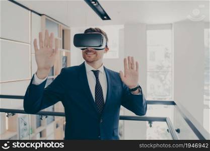 Smiling office worker in suit wearing vr headset touching air and gesturing while working in virtual reality, businessman managing business project with 3d goggles while standing in office corridor. Smiling office worker in vr headset touching air and gesturing while working in virtual reality