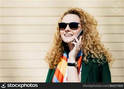 Smiling office worker in sunglasses, blonde curly hair, talking on cell phone during break. Street backdrop, beige background. Copy space available for promotional content.