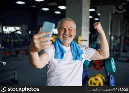 Smiling old man shows his muscles and makes selfie on phone camera, gym interior on background. Sportive grandpa on fitness training in sport center. Healty lifestyle, health care, elderly sportsman. Old man shows his muscles and makes selfie, gym