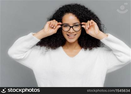 Smiling optimistic curly female with pleased facial expression, keeps hands on rim of spectacles, has bad eyesight, wears white casual jumper, models against grey studio wall. Emotions and feelings