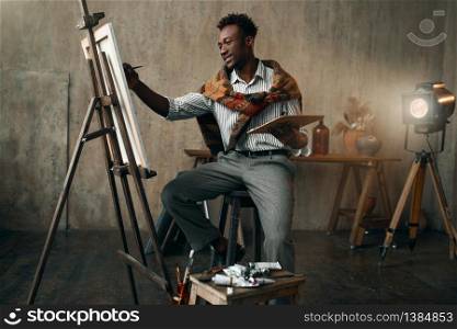 Smiling painter with brush and palette sitting at the easel. Male artist draws at his workplace, creative master works in workshop. Smiling painter with brush and palette at easel