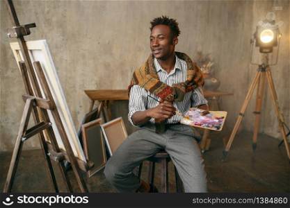 Smiling painter with brush and palette sitting at the easel. Male artist draws at his workplace. Smiling painter with brush and palette at easel